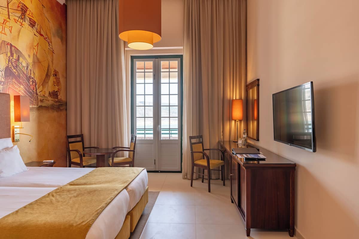 A hotel room with a bed, desk, chairs, a television, and a large window with curtains.