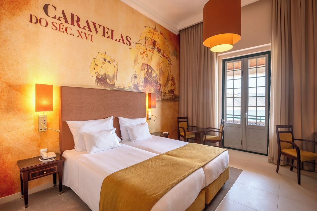 Ein Hotelzimmer mit einem großen Bett, zwei Nachttischen, einem Wandgemälde, einem Fenster mit Vorhängen und einer Sitzecke mit Stühlen.