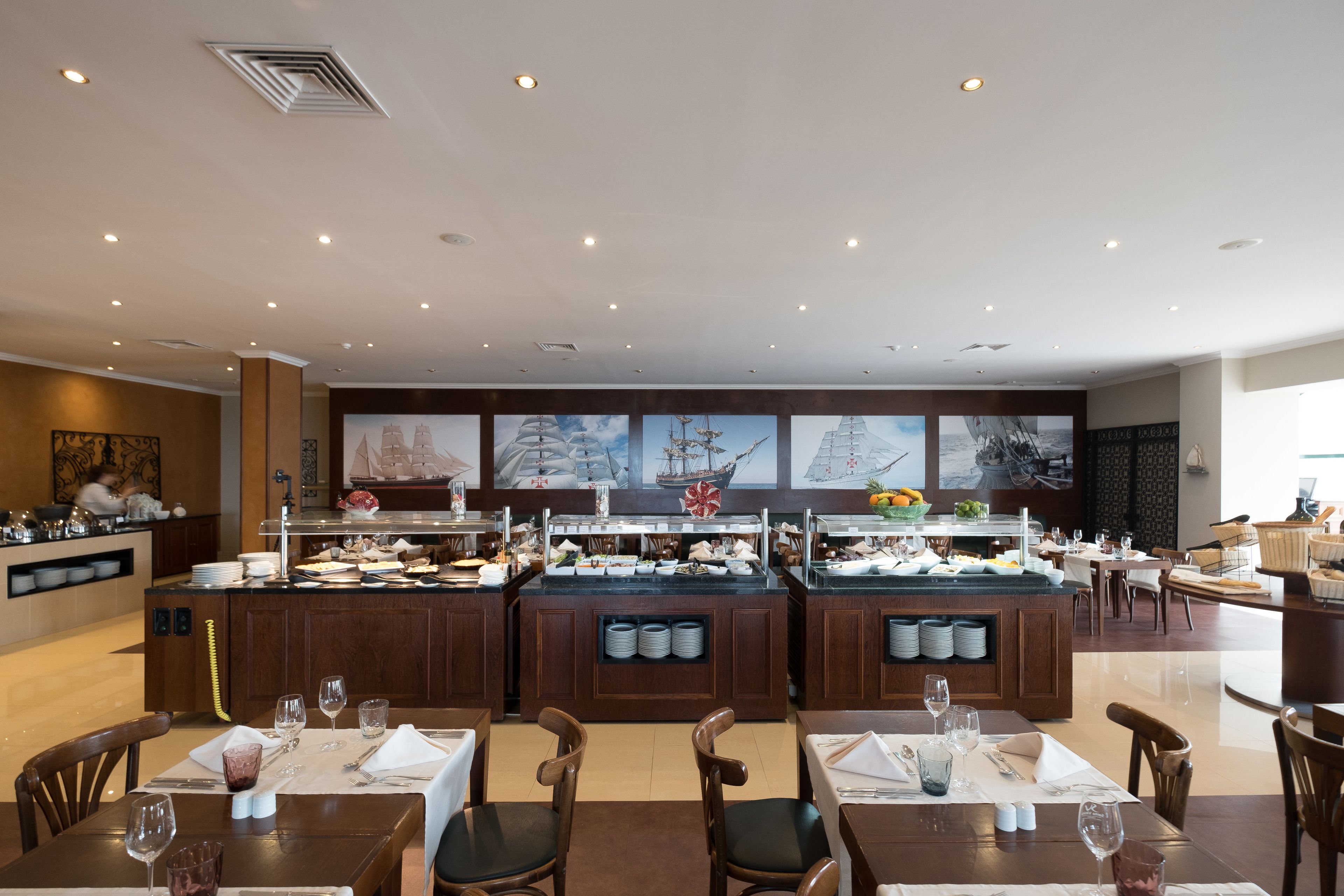 A hotel dining area with multiple buffet stations, tables set for dining, and a large mural on the wall.