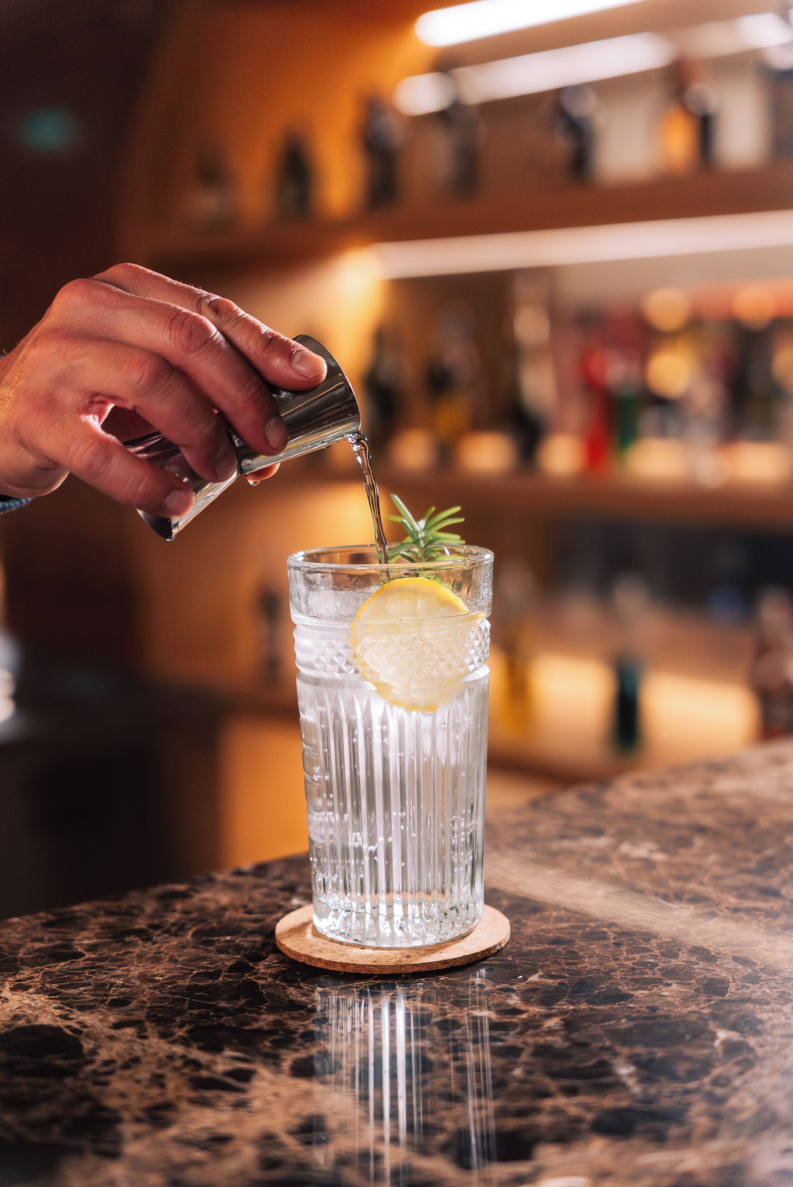 A hand pouring a drink into a glass with a lemon slice and a sprig of rosemary.