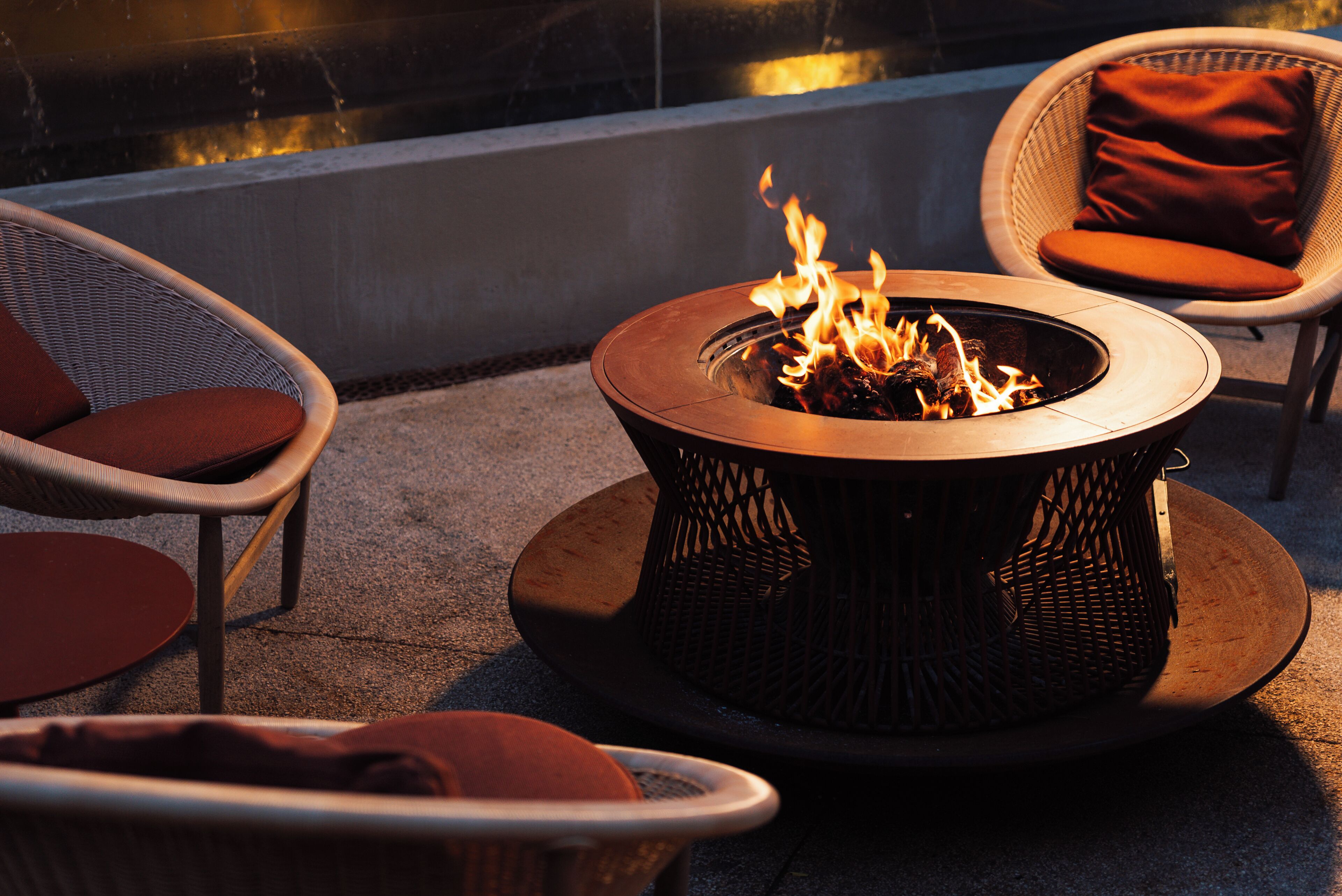 Una chimenea con llamas, rodeada de sillas de mimbre con cojines, en un patio.