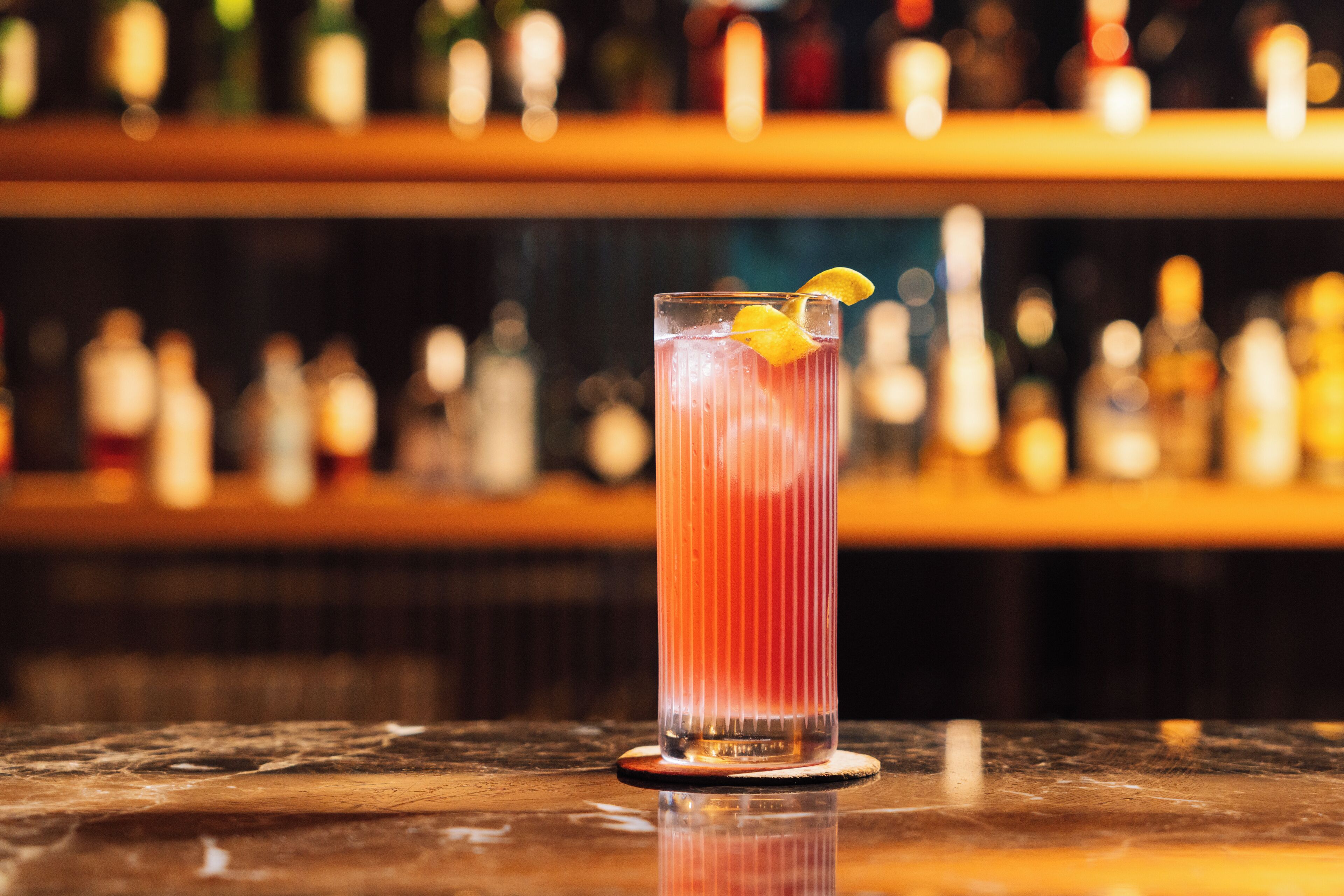 Un bar con un vaso alto de una bebida roja, un adorno de limón y un fondo desenfocado de botellas.