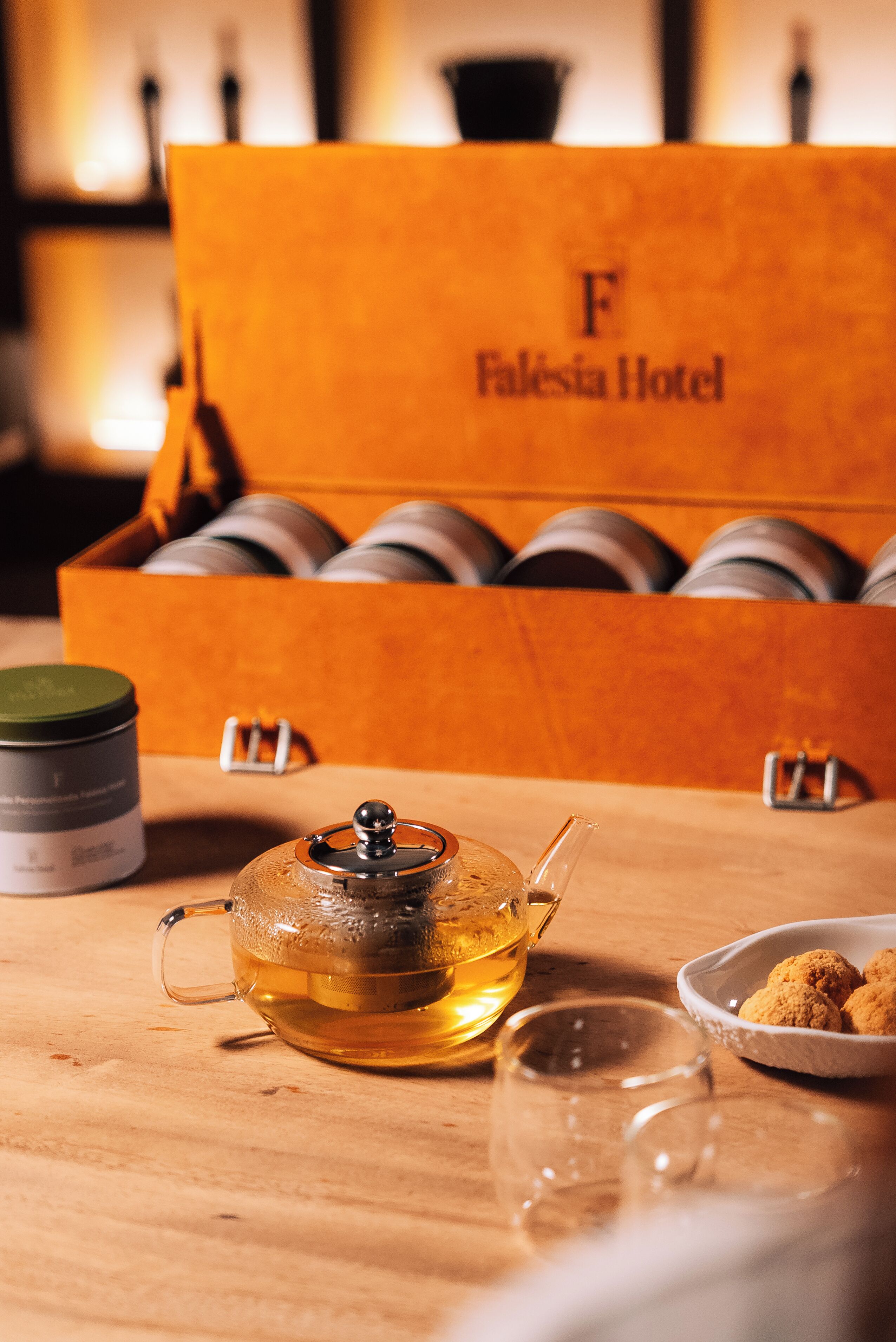 A wooden table with a teapot, a glass, and a bowl of cookies. A box labeled "Falesia Hotel" is in the background.