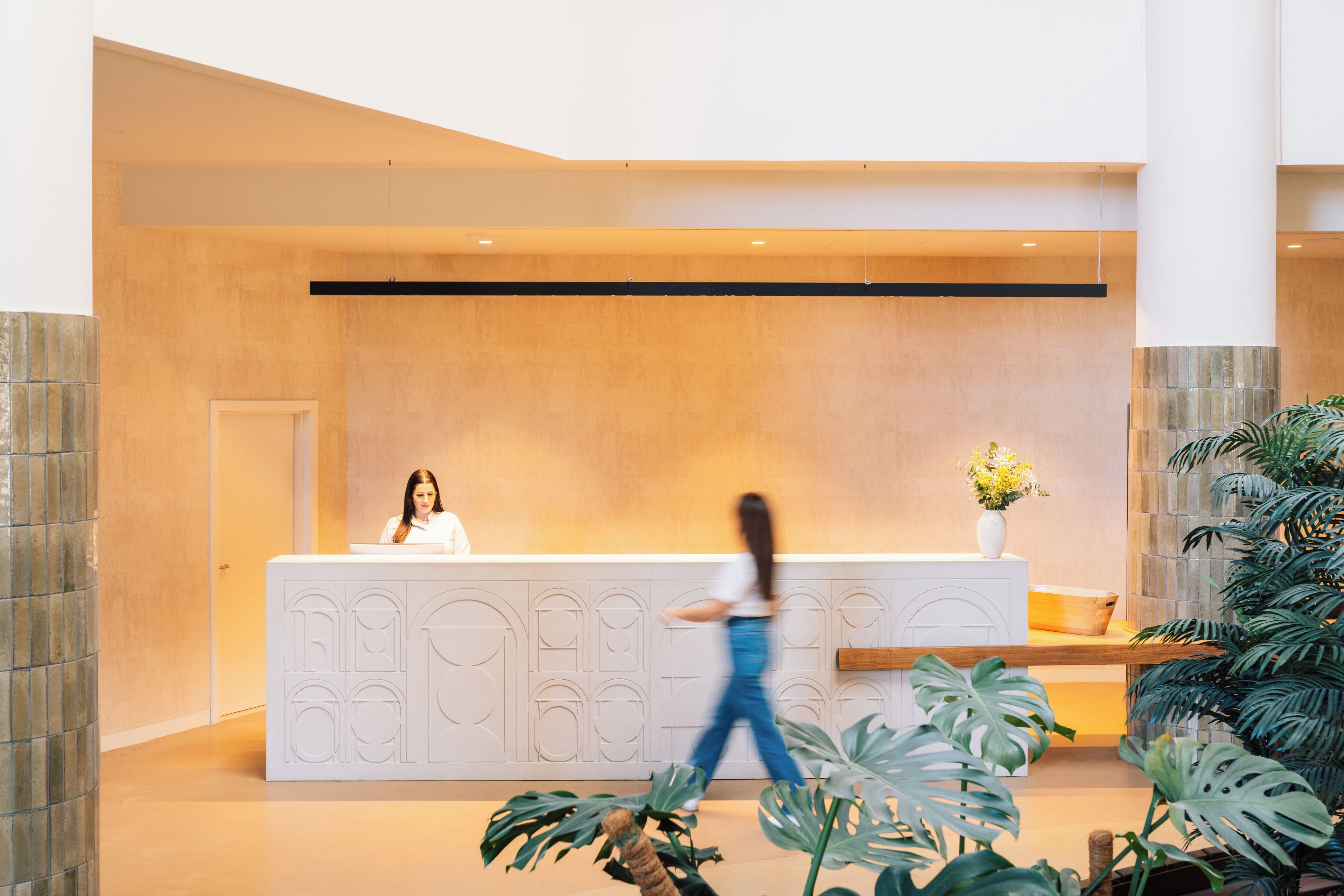 Een modern ingerichte receptie met een receptiebalie, een grote potplant en een persoon die langsloopt.