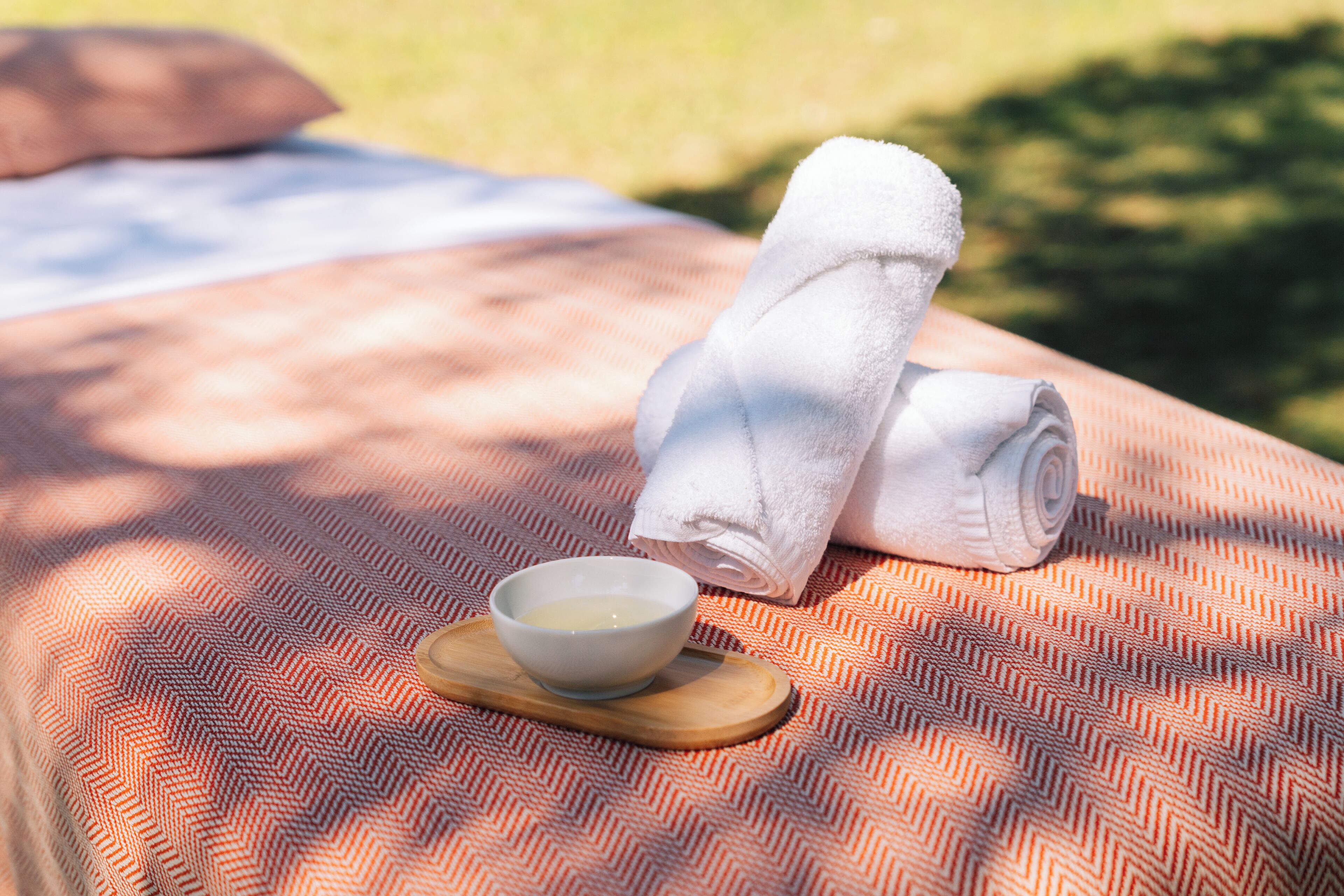 A bed with a patterned blanket, a rolled towel, and a bowl of water on a wooden tray.