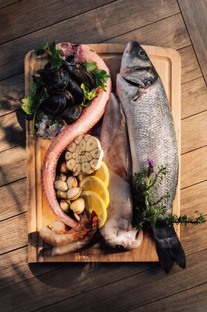 Ein Holzschnittbrett mit Meeresfrüchten, darunter Miesmuscheln, Venusmuscheln und ein ganzer Fisch, belegt mit Kräutern und Zitronenscheiben.