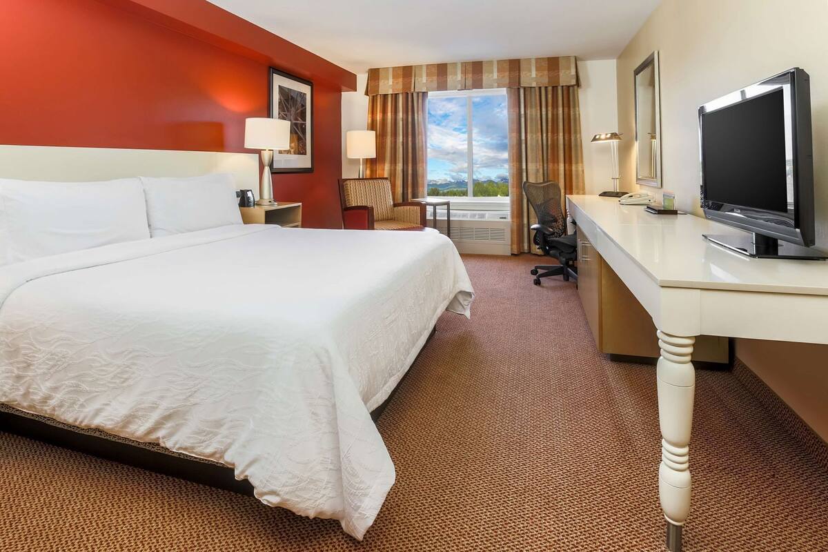 Ein Hotelzimmer mit einem großen Bett, einem Schreibtisch mit Fernseher, einem Sessel und einem Fenster mit Vorhängen.