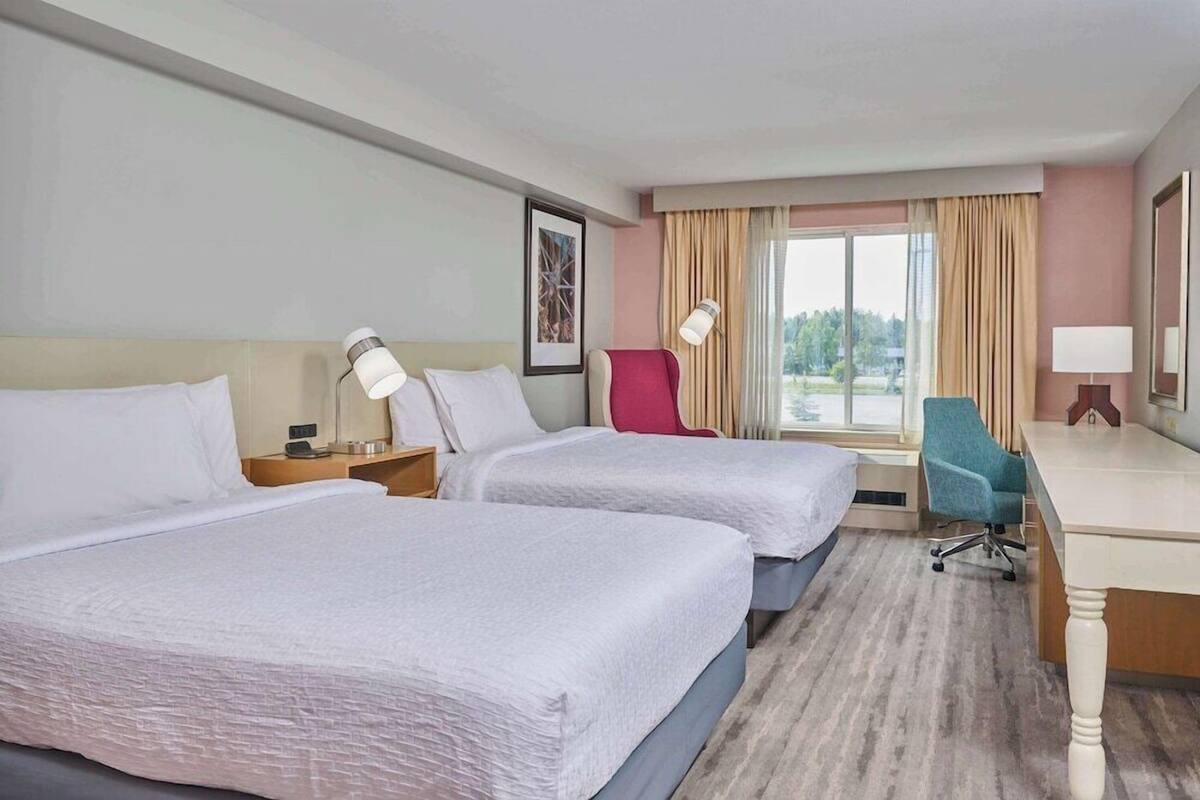 Ein Hotelzimmer mit zwei Betten, einem Schreibtisch, einem Stuhl, einer Lampe und einem fenster mit Vorhängen.