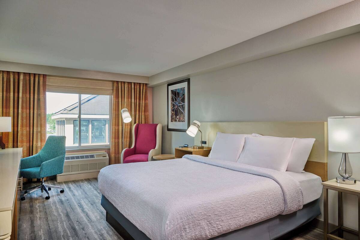 Ein Hotelzimmer mit einem großen Bett, einem Schreibtisch, einem Sessel und einem Fenster mit Vorhängen.