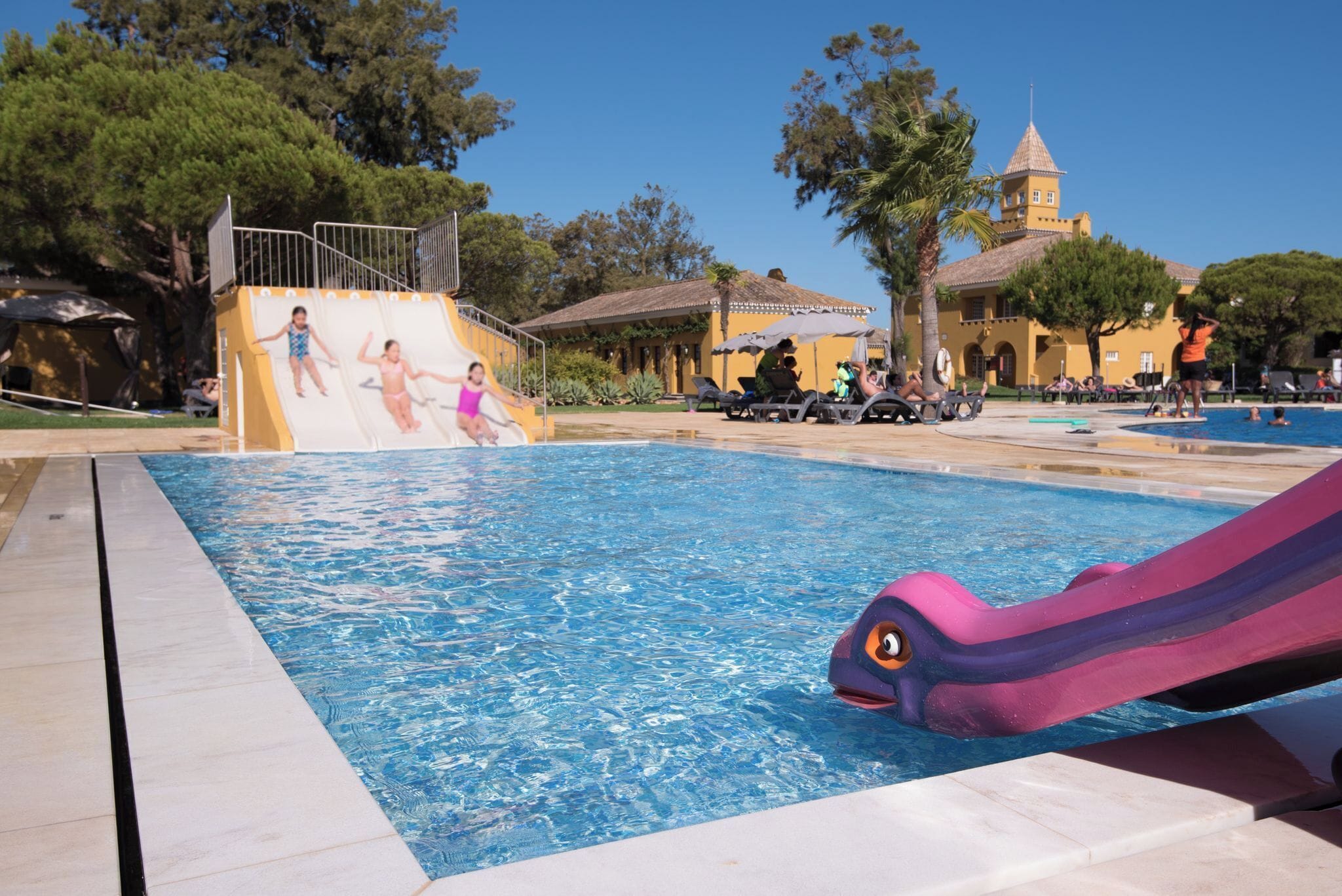 A swimming pool with a slide, a lifeguard tower, and a building with a tower in the background.