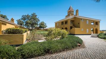 A yellow building with a tower, surrounded by greenery and a cobblestone path.