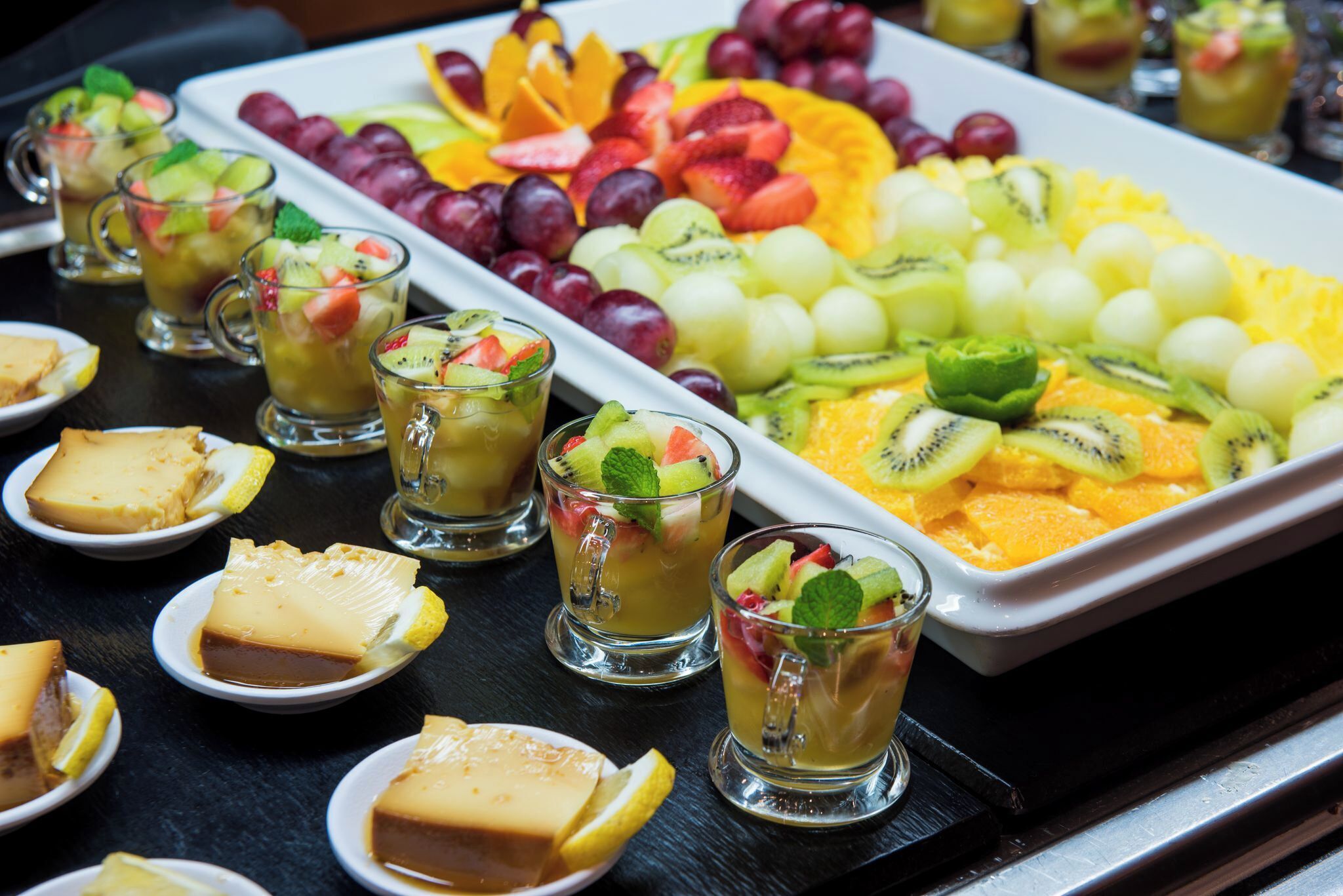 A buffet with various fruits, a tray of sliced fruits, and several glasses of a fruit drink.