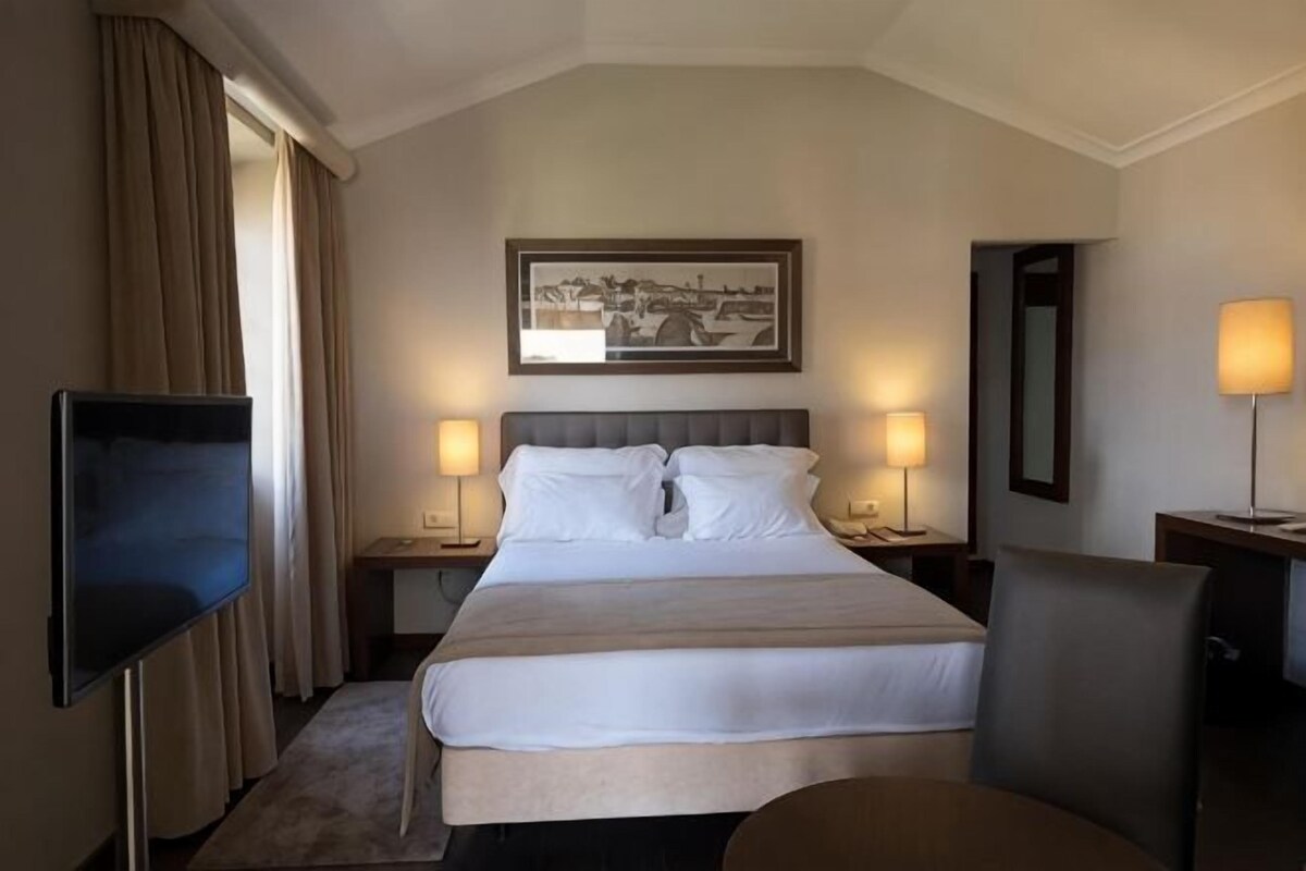 Een hotelkamer met een groot bed, twee nachtkastjes met lampen, een flatscreen televisie, een stoel en een ingelijst schilderij aan de muur.