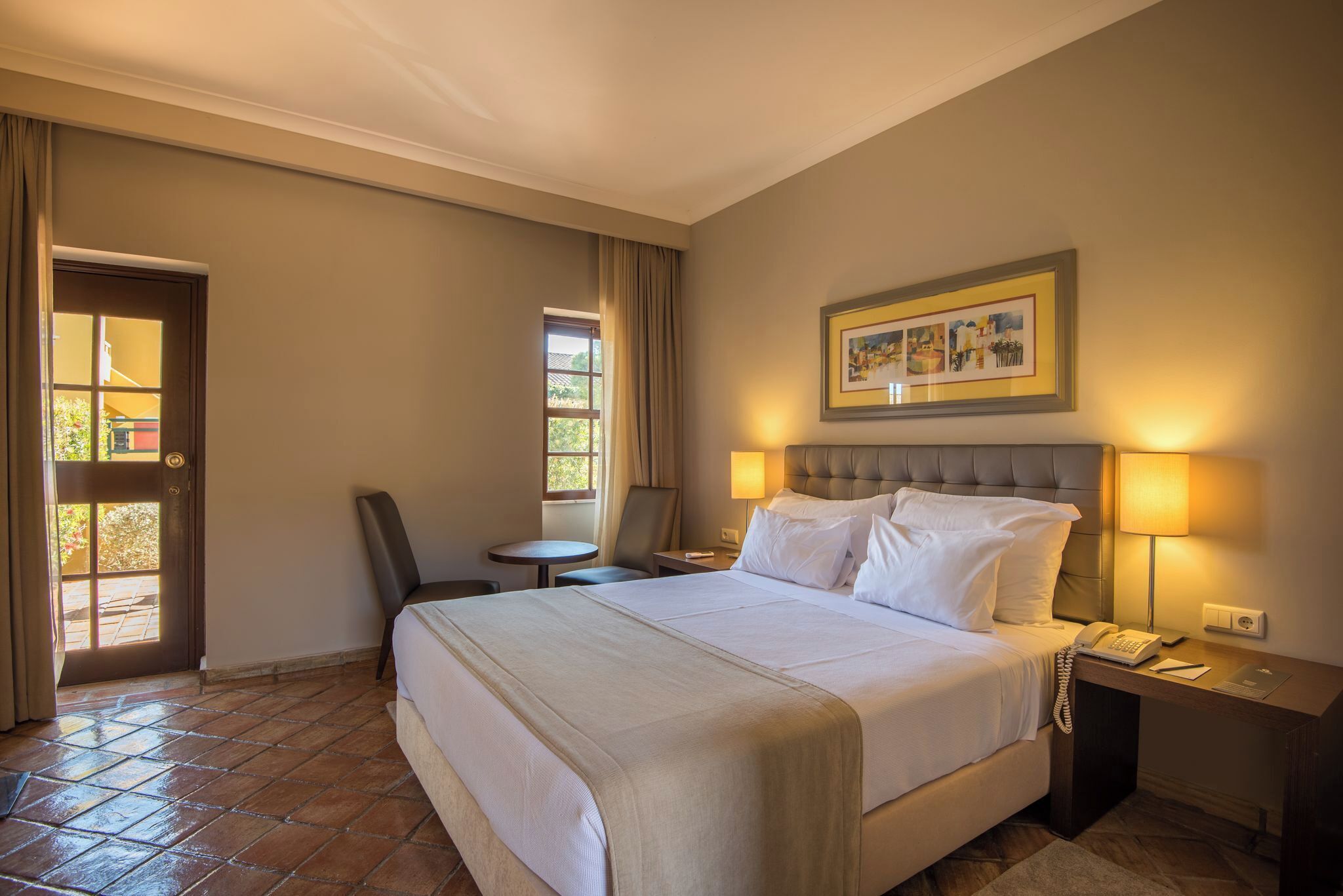 Een hotelkamer met een bed, bedlampen, een tafeltje, een stoel en een ingelijst schilderij aan de muur.