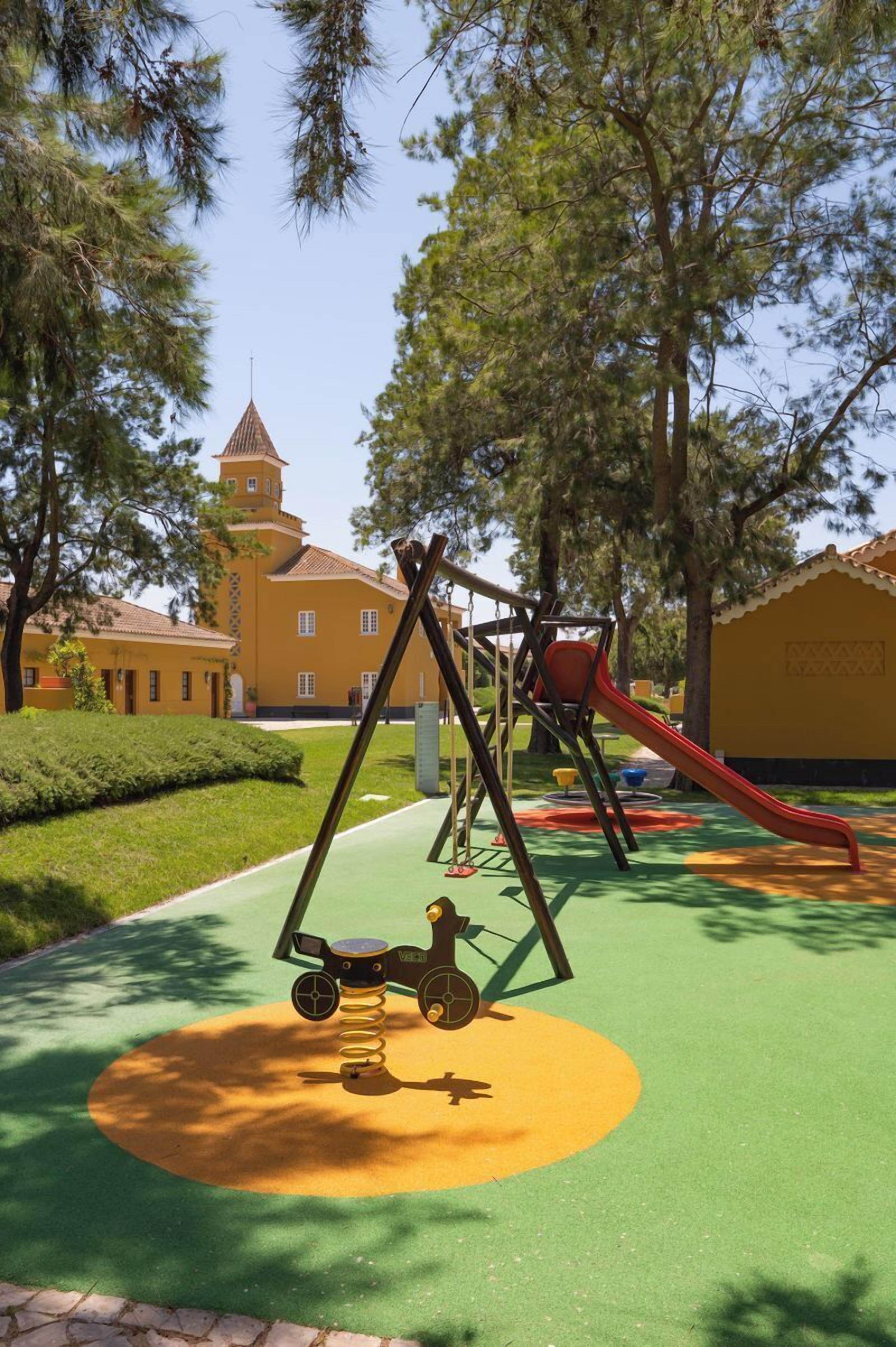 A playground with swings and a slide, surrounded by trees and a building with a tower.