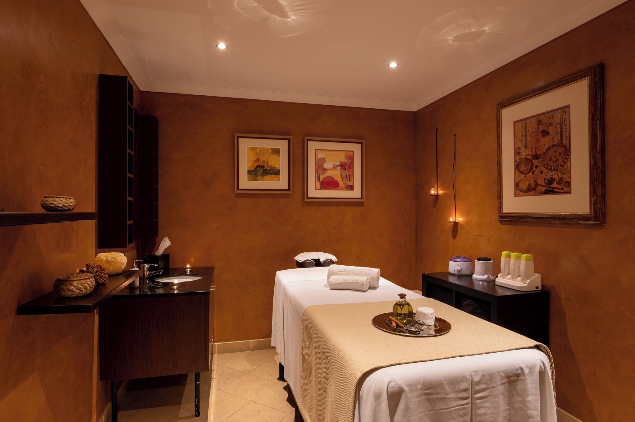 Ein Spa-Zimmer mit Massageliege, einem kleinen Schreibtisch mit Korb, einer Ablage mit Flasche und gerahmten Bildern an den Wänden.