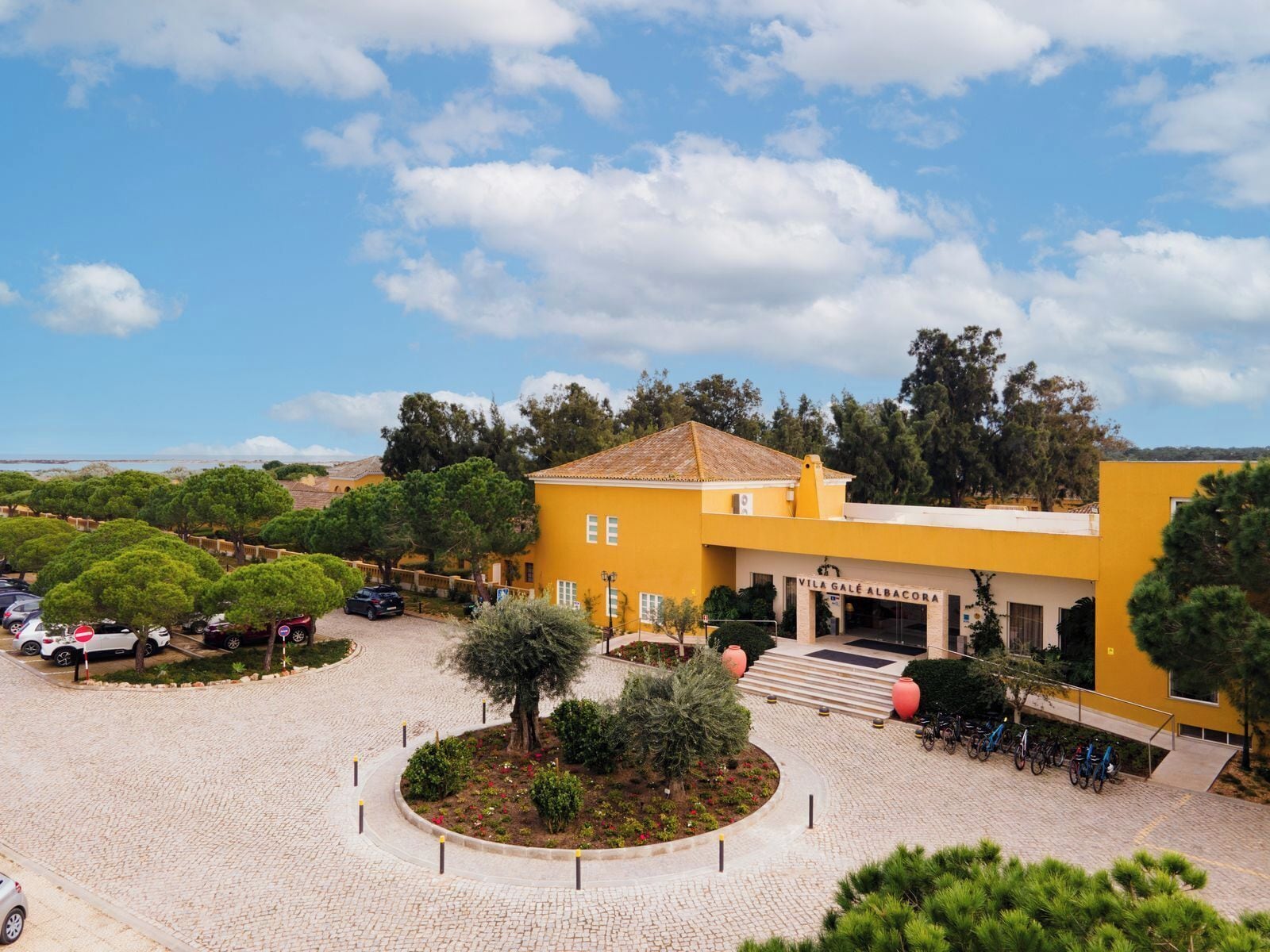 A yellow building with a sign reading 'VILA GALÉ ALBACORA,' a paved courtyard with trees, and parked bicycles.