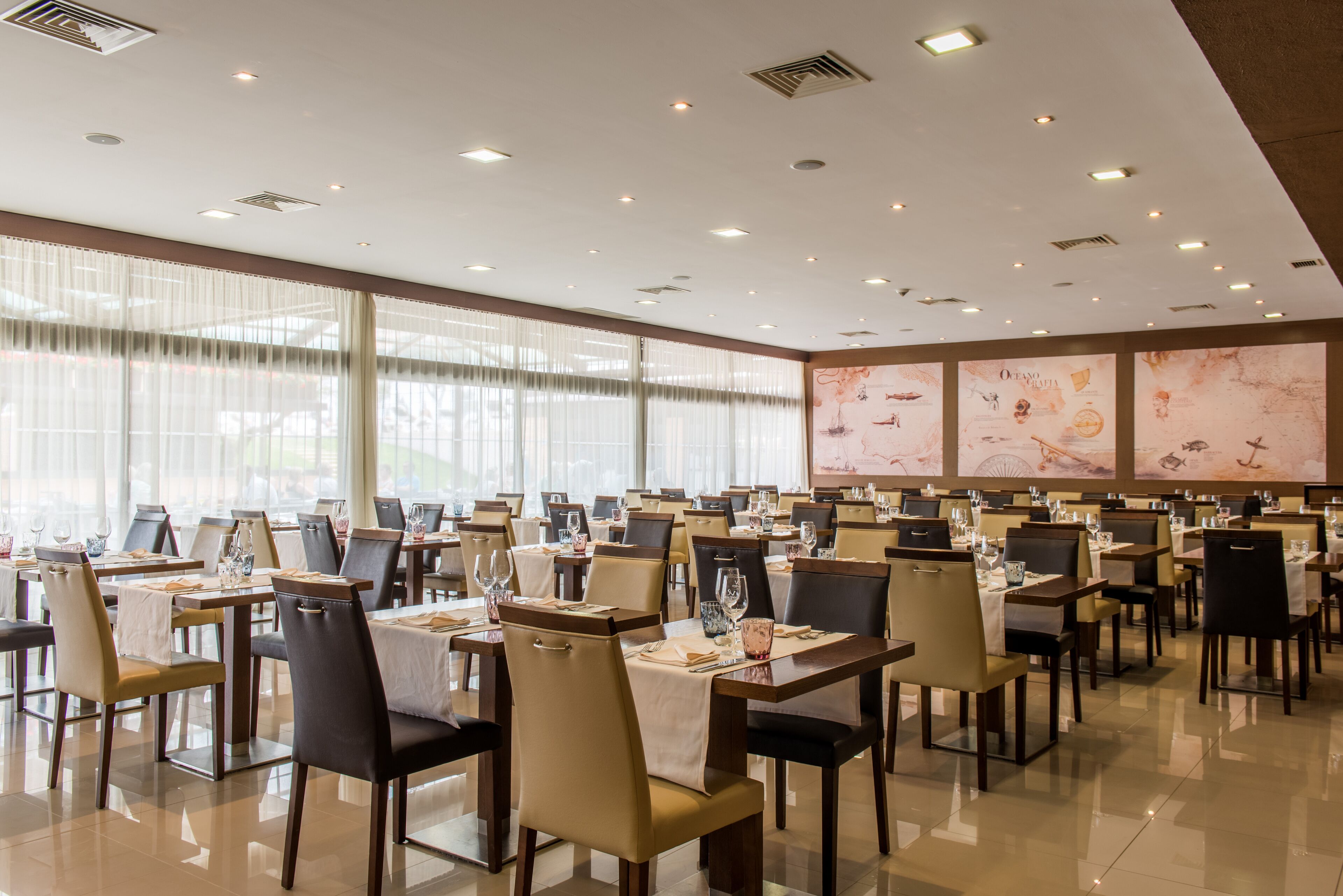 A spacious dining area with multiple tables and chairs, large windows, and ceiling lights.