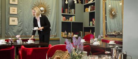 Un comedor con sillas tapizadas en rojo, una mesa de buffet con pan y un jarrón de flores, y una mesa lista para el desayuno con huevos y zumo de naranja.