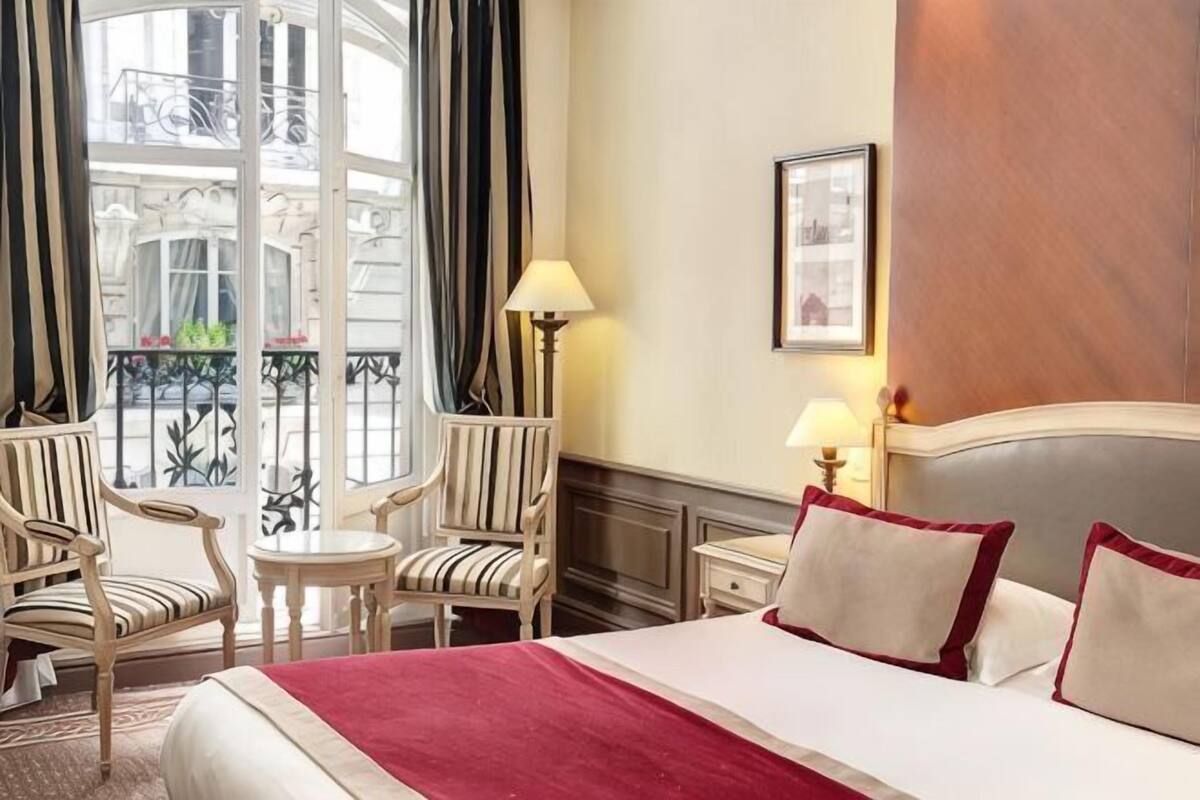 Une chambre d’hôtel avec un lit, deux tables de chevet, une lampe, une chaise, une petite table et une vue sur un balcon.