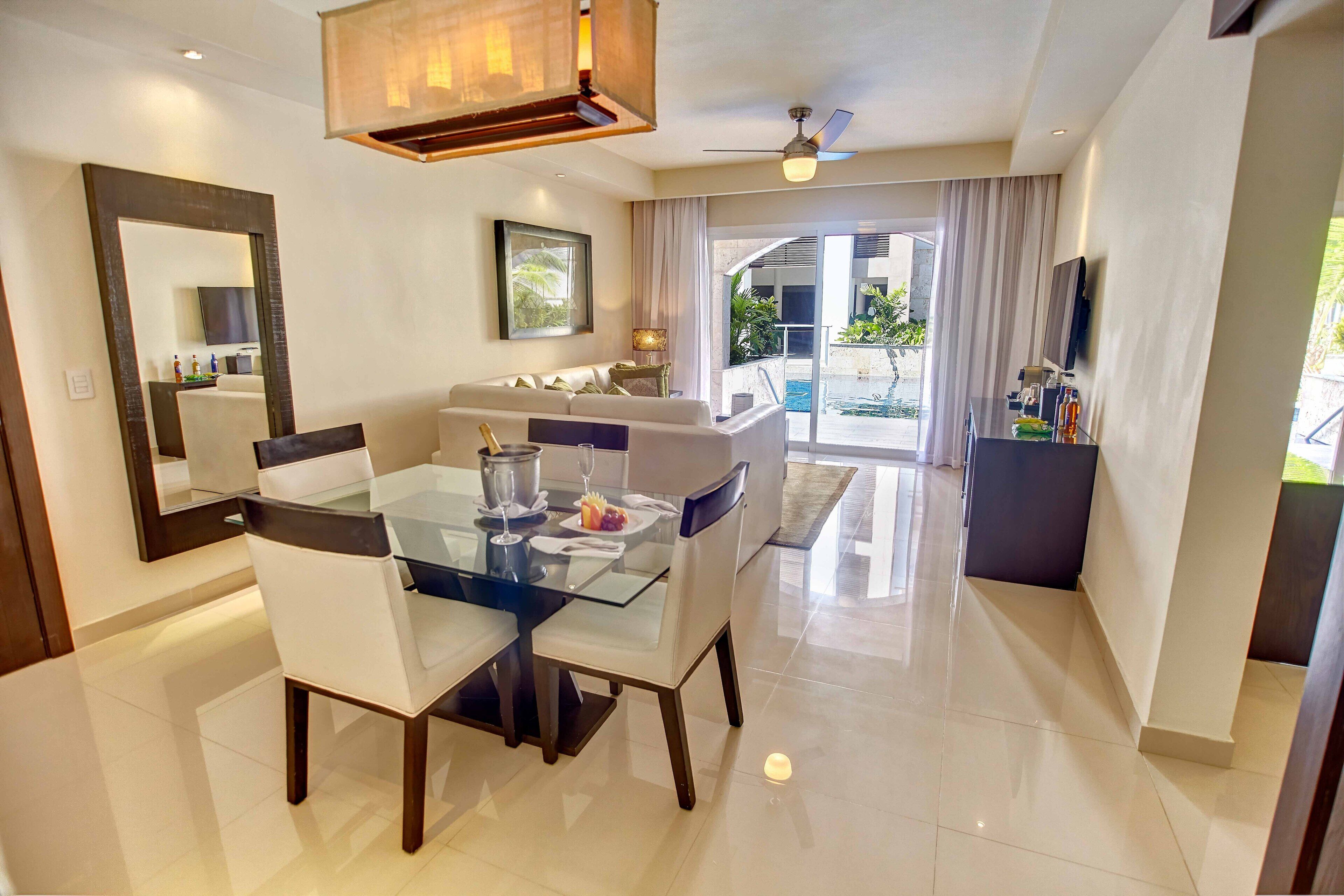 A modern hotel room with a dining area, a large mirror, a ceiling fan, and a view of the outdoor pool area.