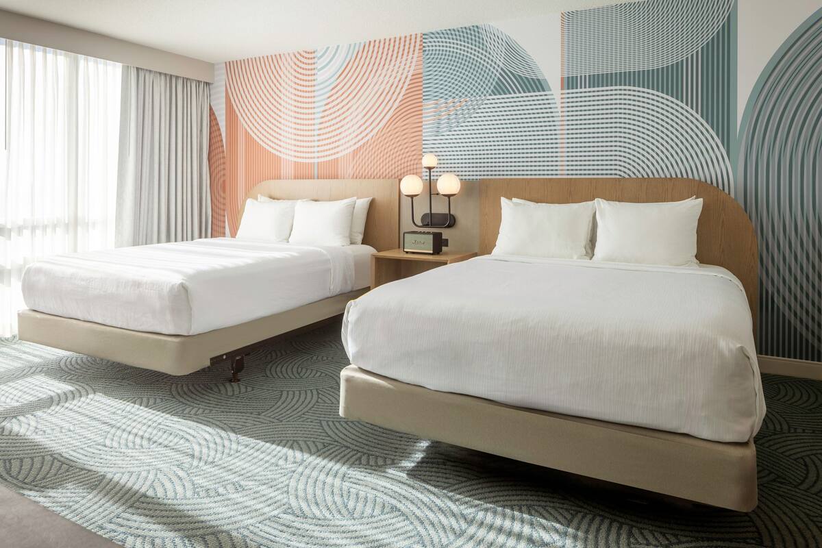 Habitación de hotel con dos camas, una mesita de noche con lámpara y un mural abstracto grande en la pared.