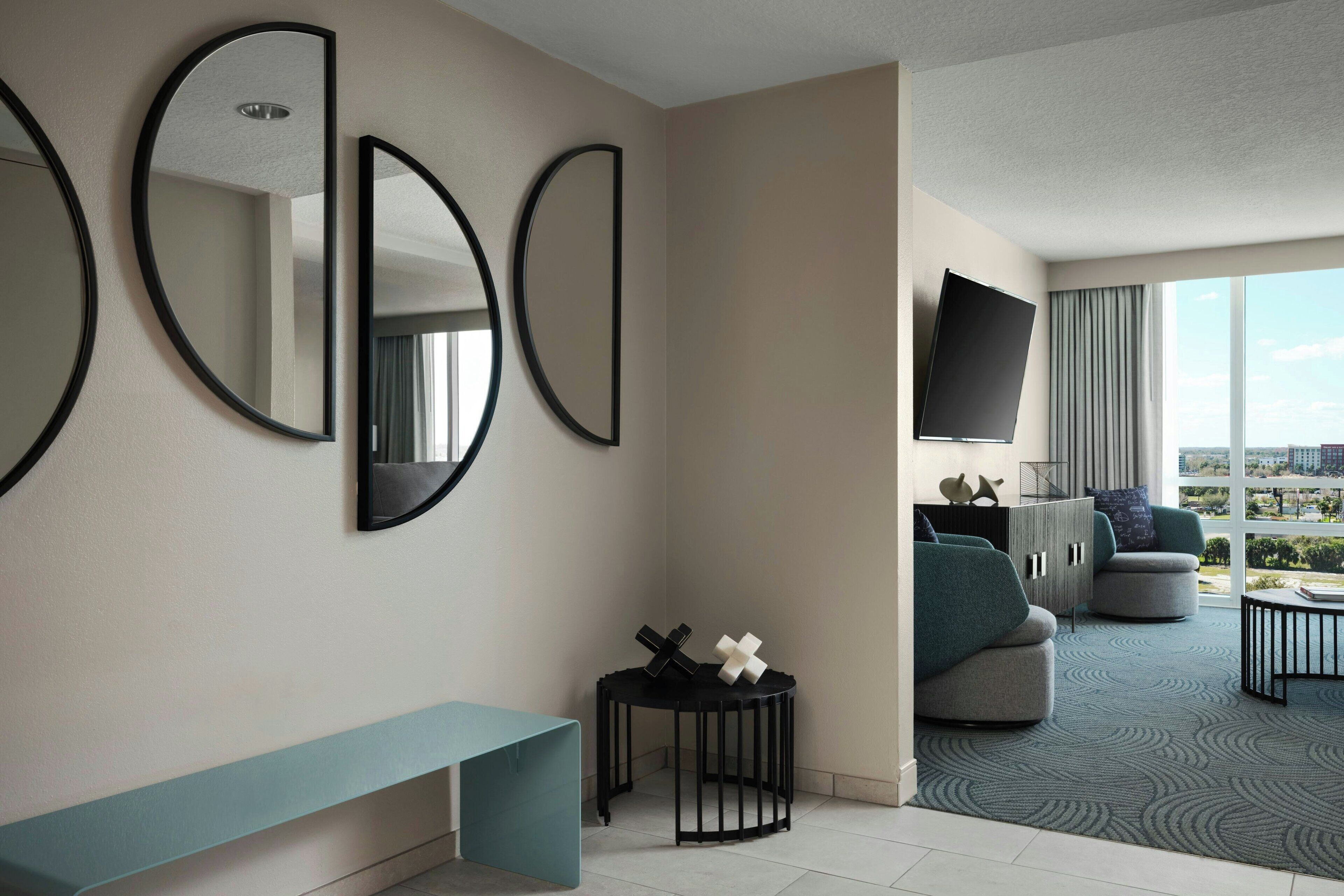 A modern hotel room with a large window, a flat-screen TV, a sofa, a coffee table, and a round side table with decorative items.