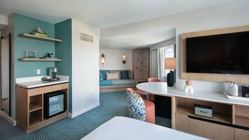 A hotel room with a bed, a desk with a refrigerator, a TV, a seating area with a sofa, and a small table with a lamp.
