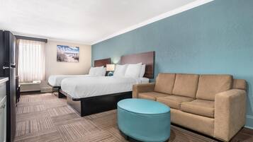 Habitación de hotel con cama, dos mesitas de noche, sofá, pequeño reposapiés y televisión instalada en la pared.