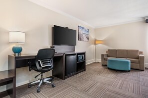 Et hotelværelse med skrivebord, stol, fjernsyn, sofa, skammel, lampe og et billede på væggen.