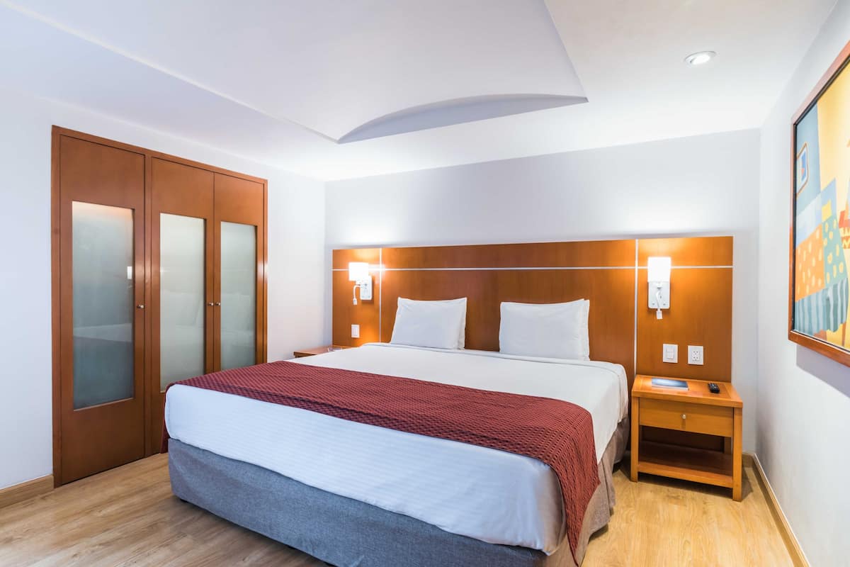 A hotel room with a large bed, wooden wardrobe, bedside tables, and a wall-mounted light fixture.