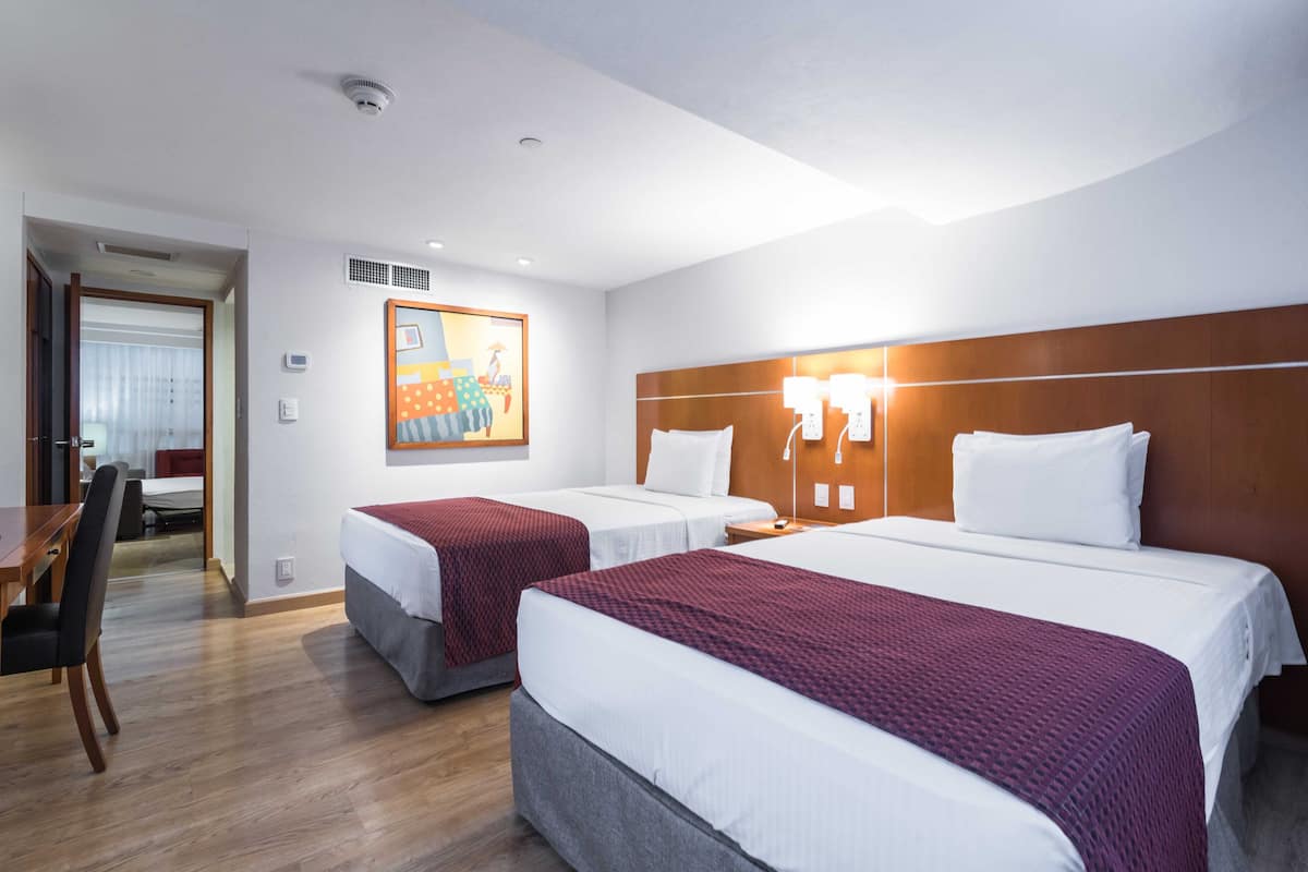 A hotel room with two beds, a wooden headboard, a painting on the wall, and a desk with a chair.