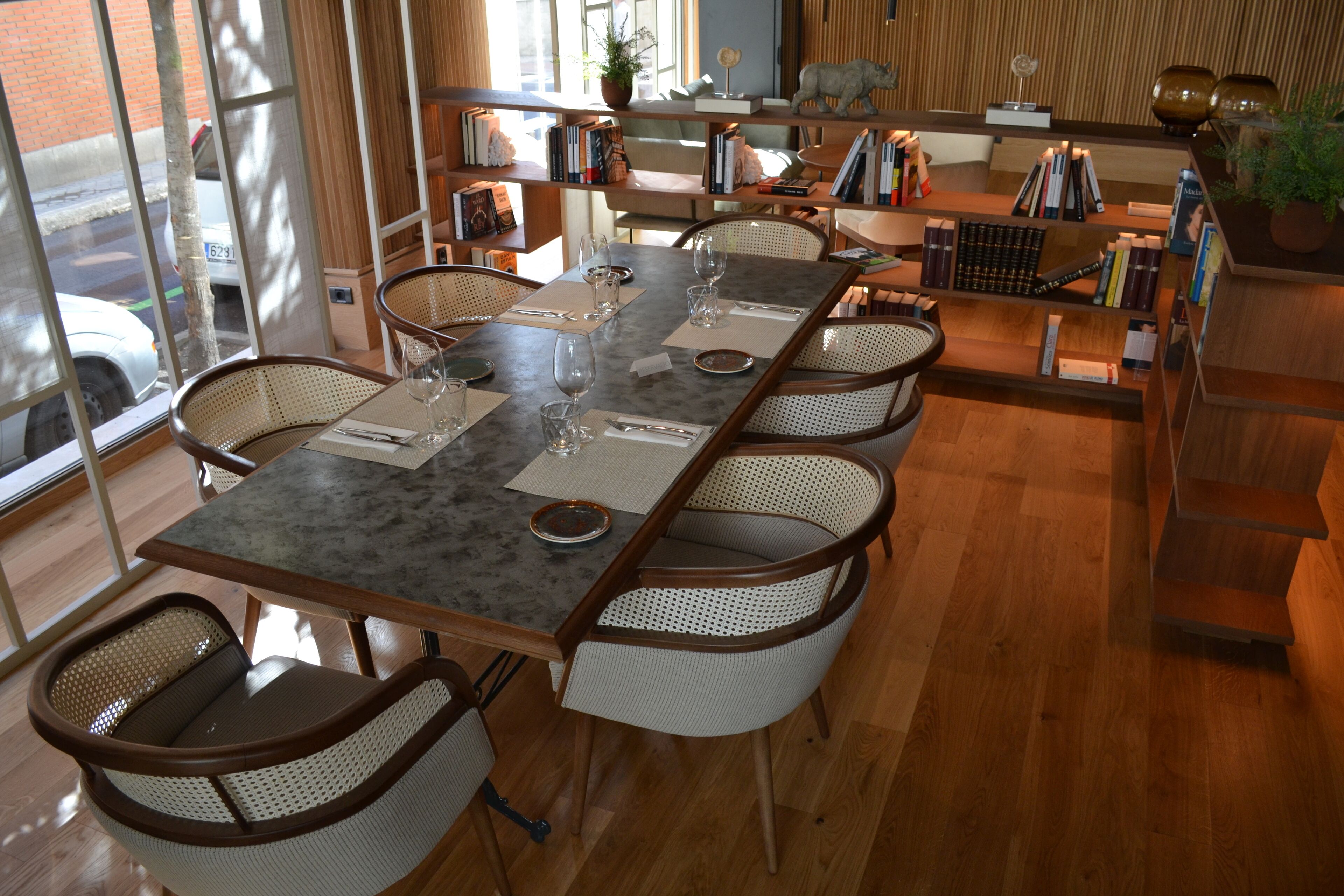 A dining area with a long table, six wicker chairs, and a bookshelf in the background.