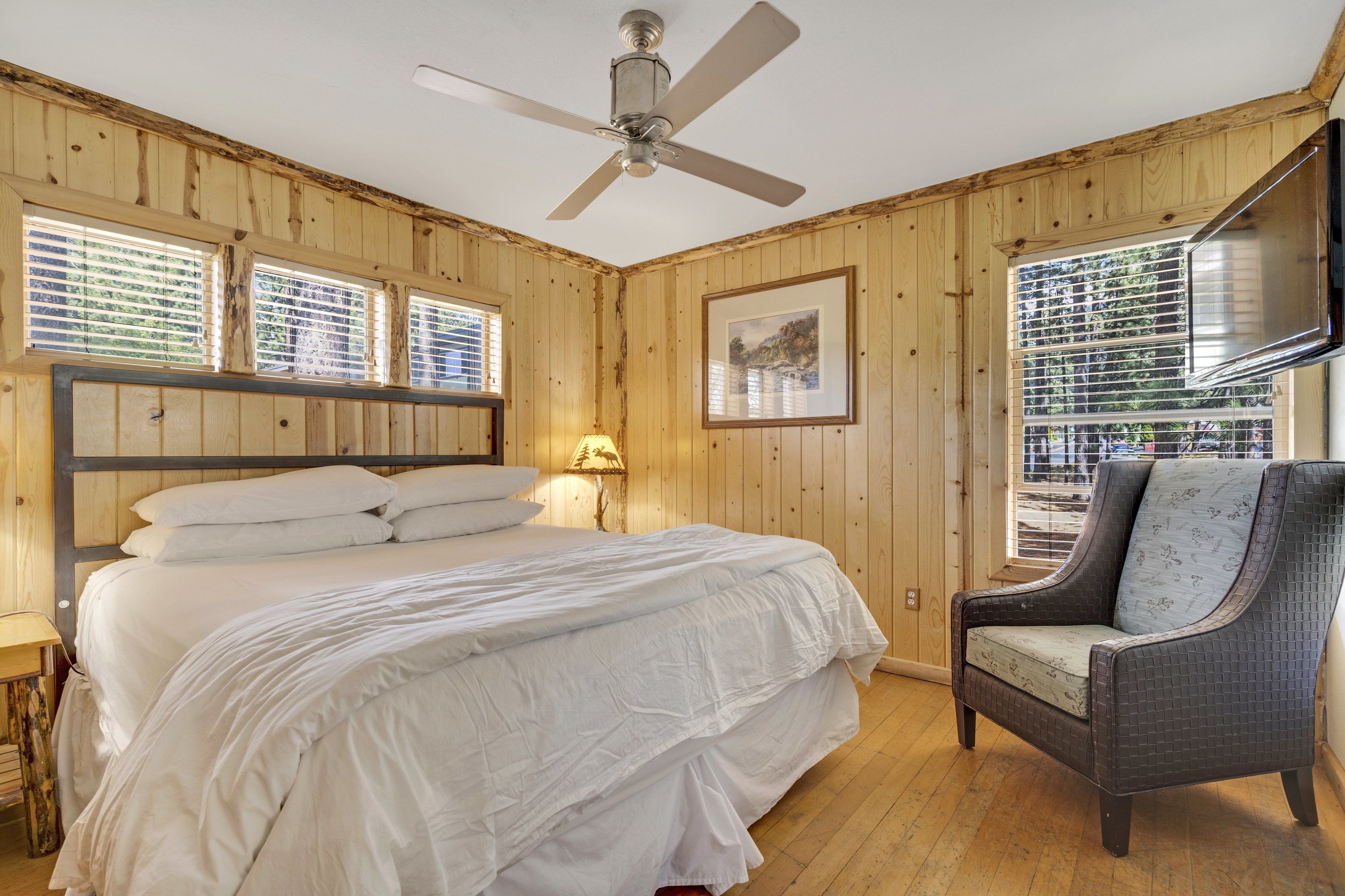 A bedroom with a bed, a chair, a ceiling fan, wooden walls, and a window with blinds.