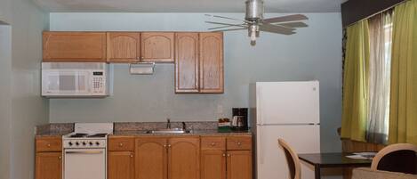 A compact kitchen with wooden cabinets, a microwave, a stove, a refrigerator, and a ceiling fan.
