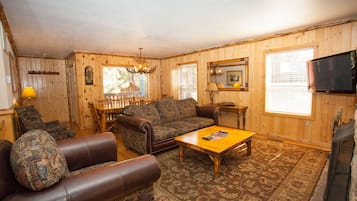 A cozy living room with a brown leather sofa, patterned armchair, and a wooden coffee table. A television is mounted on the wall, and a fireplace is visible in the corner.