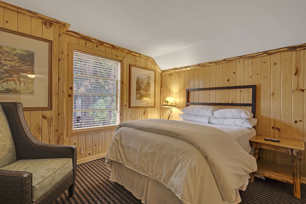 A wooden-paneled room with a bed, a chair, a window with blinds, and framed pictures on the wall.