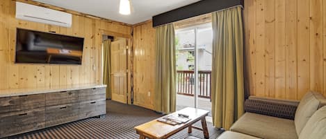 Una habitación con interiores de madera, un ventilador de techo, un televisor de pantalla plana montado en la pared, un sofá, una mesita y un ventanal corredero que da a un espacio exterior.