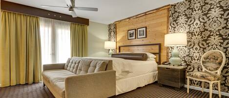 Habitación de hotel con cama, sofá, sillón, mesita de noche, lámpara y ventilador de techo.