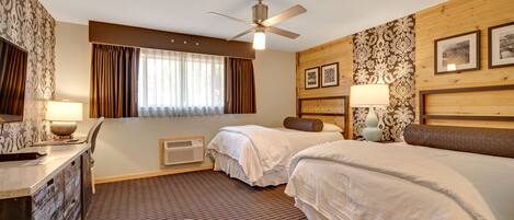 A hotel room with two beds, a ceiling fan, a desk with a lamp, a chair, and a window with curtains.