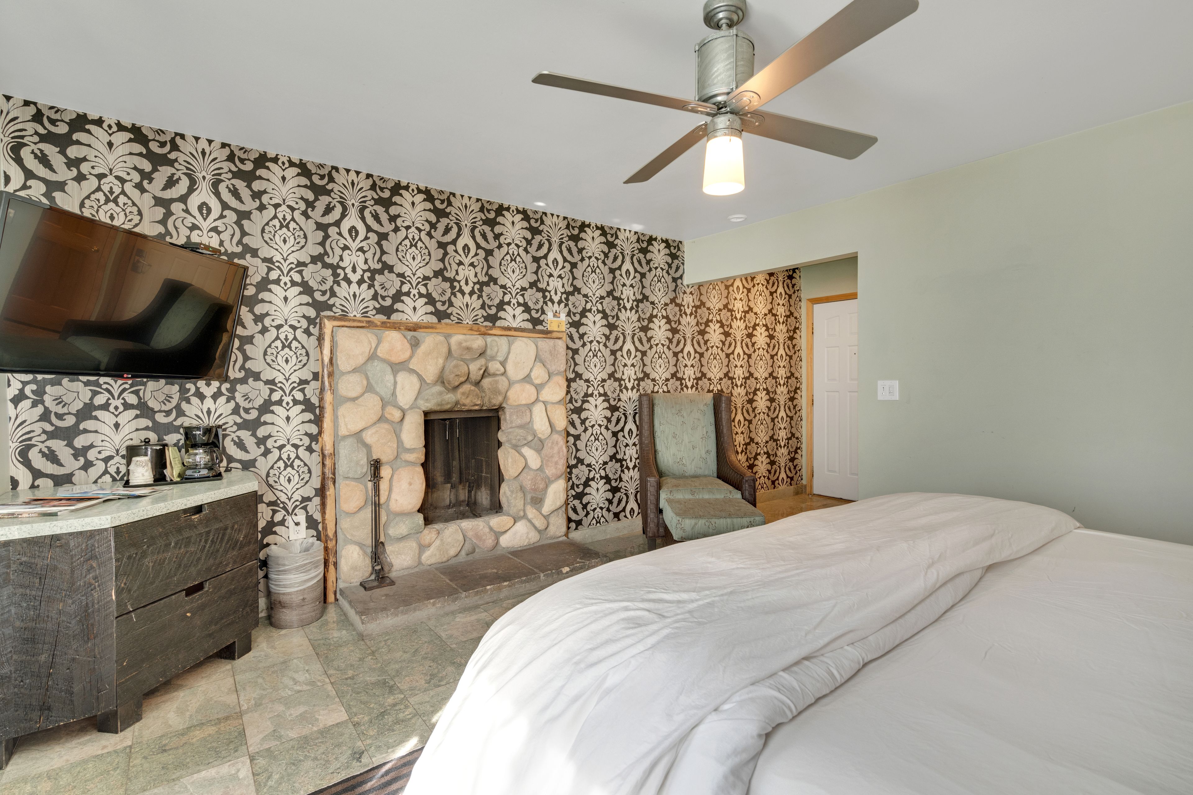 A bedroom with a stone fireplace, a ceiling fan, a flat-screen TV, and a dresser.