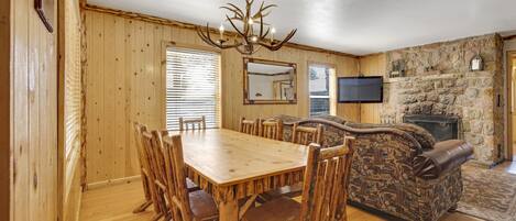 A wooden dining table with chairs, a stone fireplace, a television, and a patterned sofa in a cabin-style room.