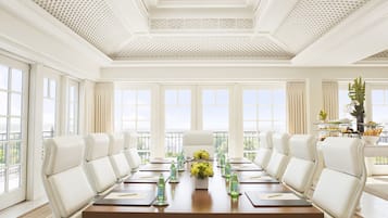 A conference room with a long table, white chairs, and a view of the outdoors through large windows.