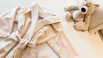 Una cama ordenada con edredón blanco, bata color beige, zapatillas y un peluche.