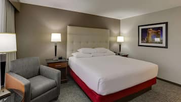 Habitación de hotel con una cama grande, un sillón gris, dos mesitas de noche con lámparas y un cuadro en la pared.