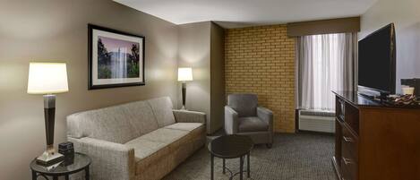 Habitación de hotel con sofá, sillón, mesita redonda, televisión y una pared de acento de ladrillo.