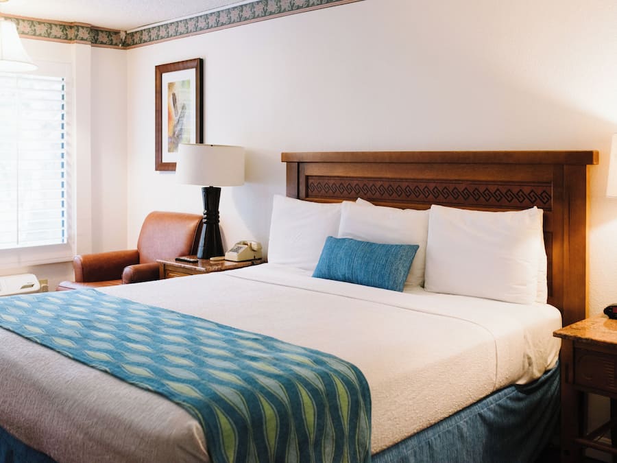 A hotel room with a large bed, a nightstand with a lamp, a chair, and a wall clock.