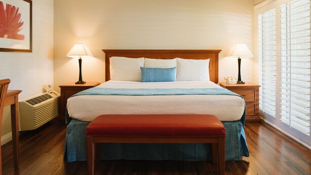 A neatly arranged hotel room with a large bed, two bedside tables, a bench, a heater, and a window with blinds. - Alamo Inn & Suites (Anaheim)