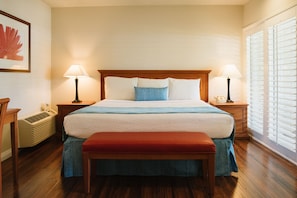 A neatly arranged hotel room with a large bed, two bedside tables, a bench, a heater, and a window with blinds.