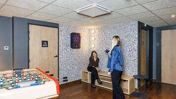 A room with a foosball table, a dartboard, and a seating area with two people.